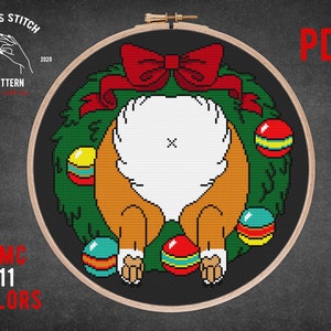 May include: Cross-stitch pattern featuring a dog's rear end inside a Christmas wreath with a red bow. The design includes colorful ornaments and the text "PDF", "DMC 11 COLORS", and "CROSS STITCH PATTERN". The pattern is set in a wooden embroidery hoop.