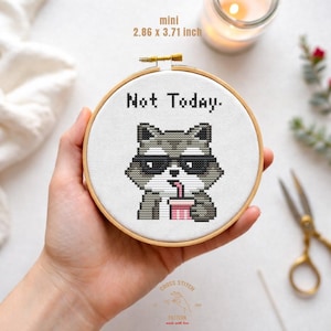 May include: Miniature cross-stitch embroidery in a wooden hoop. The design features a pixelated raccoon wearing sunglasses and drinking from a pink cup, with the words "Not Today." The hoop measures 7.26 x 9.42 cm.