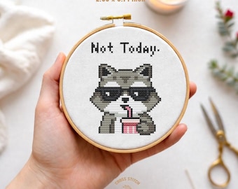 Pode incluir: Miniatura de bordado em ponto cruz em um bastidor de madeira. O desenho apresenta um guaxinim pixelado usando óculos de sol e bebendo de uma xícara rosa, com as palavras "Not Today". O bastidor mede 7,26 x 9,42 cm.