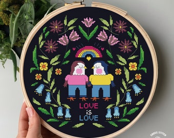 May include: Cross stitch pattern featuring two birds holding hands in front of a rainbow with the text "Love is Love" surrounded by flowers and leaves on a dark blue background.