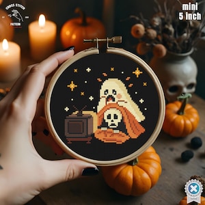 May include: A mini 5-inch cross-stitch embroidery hoop featuring a Halloween design. The design includes a ghost, a skull, a pumpkin, and a vintage television set, all stitched on a black background with star accents. The hoop is held by a hand.