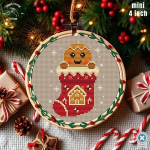 May include: A mini 4-inch cross-stitch Christmas ornament featuring a gingerbread man in a red stocking. The design is framed in a wooden hoop with green leaves and red berries. The background is a festive holiday scene.