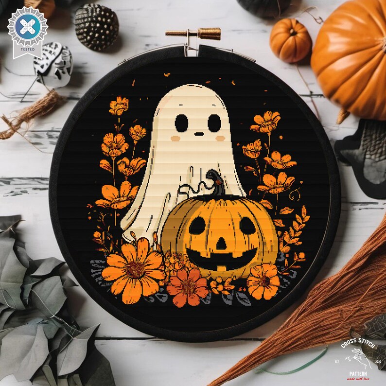 May include: A cross-stitch design in a black hoop, featuring a ghost and a carved pumpkin. The design is surrounded by orange flowers. The hoop is displayed on a white wooden surface, with pumpkins and other decorations.