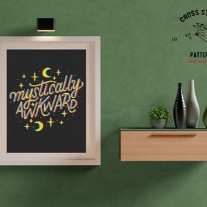 May include: A framed cross stitch pattern with the text "mystically awkward" in rainbow colors with stars and crescent moons. The pattern is on a black background and is framed in white. The frame is hanging on a green wall.