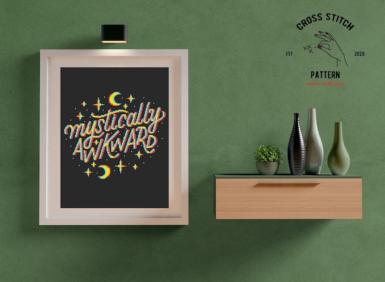 May include: A framed cross stitch pattern with the text "mystically awkward" in rainbow colors with stars and crescent moons. The pattern is on a black background and is framed in white. The frame is hanging on a green wall.