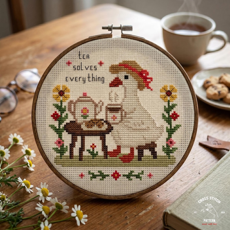 May include: A cross-stitch embroidery in a wooden hoop features a goose enjoying tea. The design includes a teapot, flowers, and the text "Tea Solves Everything." The color palette includes beige, brown, red, yellow, and green.