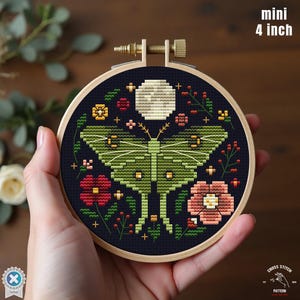 May include: A cross-stitch embroidery hoop featuring a green moth design with a full moon and floral accents. The embroidery is set in a 4-inch (10 cm) wooden hoop, with the text "mini 4 inch" visible.