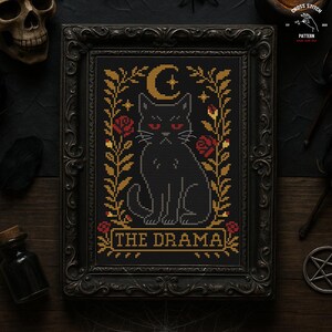 May include: Cross-stitch pattern in a black ornate frame. The design features a black cat with red eyes, a crescent moon, stars, and roses. The text "THE DRAMA" is at the bottom. The overall theme is gothic and spooky.
