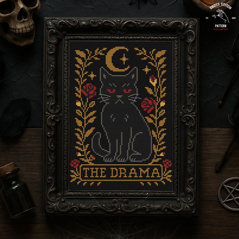 May include: Cross-stitch pattern in a black ornate frame. The design features a black cat with red eyes, a crescent moon, stars, and roses. The text "THE DRAMA" is at the bottom. The overall theme is gothic and spooky.