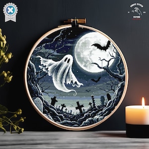 May include: A cross stitch pattern featuring a ghost flying in front of a full moon with bats and a graveyard in the foreground. The pattern is framed in a round wooden hoop.