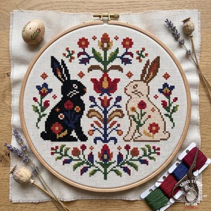 Op de afbeelding: Een ronde houten borduurring met een kruissteekpatroon op gebroken wit stof. Het ontwerp toont een zwart konijn en een crèmekleurig konijn tegenover een centraal bloemmotief, omringd door kleine rode, blauwe en groene bloemaccenten.