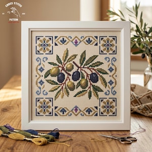 May include: A framed cross-stitch pattern featuring an olive branch with green and dark purple olives. The design is set against a beige background with a decorative border in blue, gold, and purple. The frame is white, and embroidery floss and scissors are visible.