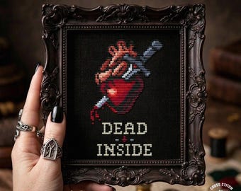 Pode incluir: Uma peça de ponto cruz emoldurada com uma estética sombria. A obra de arte apresenta um coração vermelho perfurado por uma adaga prateada, com as palavras "DEAD INSIDE" bordadas abaixo. A moldura é ornamentada e castanho escuro, e a peça mede 8 x 12.7 cm.