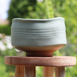 May include: A light green ceramic tea bowl with a slightly uneven rim and a textured surface. The bowl is sitting on a wooden stool.