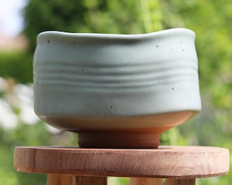May include: A light green ceramic tea bowl with a slightly uneven rim and a textured surface. The bowl is sitting on a wooden stool.