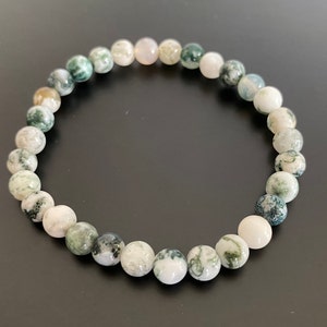 Op de afbeelding: Een armband van ronde, gepolijste stenen. De stenen zijn voornamelijk wit met groene en grijze insluitsels, wat een natuurlijk patroon creëert. De armband wordt getoond op een donkere ondergrond.