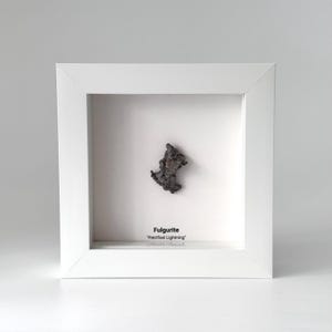 May include: A dark, textured, irregular fulgurite specimen displayed inside a square white frame. The piece is centred against a white background with the text "Fulgurite" and "Petrified Lightning" printed in black below the specimen.
