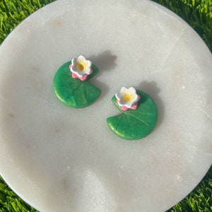 Peut inclure: Une paire de boucles d'oreilles en forme de nénuphar vert et blanc avec une fleur rose au centre. Les boucles d'oreilles sont sur une surface en marbre blanc.