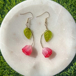 May include: A pair of gold earrings with pink flower and green leaf charms. The earrings are on a white marble surface.