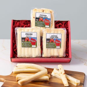 May include: A red gift box filled with red paper and three packages of string cheese. The packages are labeled "Pepper String Cheese", "String Cheese", and "Smoked String Cheese" from the Wisconsin Cheese Company.