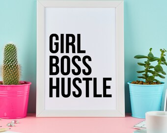 以下が含まれることがあります： 白い額縁に入ったプリントで、黒い文字で「GIRL BOSS HUSTLE」と書かれています。プリントはピンクの表面にあり、ピンクの鉢植えの仙人掌と青い鉢植えの多肉植物があります。