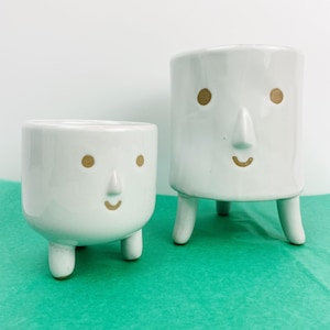 May include: Two white ceramic plant pots with cartoon faces. The pots have three legs and gold-coloured eyes and smiles. One pot is taller than the other. The background is white and the surface is teal.