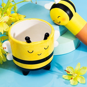 May include: A yellow ceramic bee-themed planter with black stripes and a smiling face. The planter has white wings and black antennae. A matching bee-shaped item is also visible, along with yellow flowers, all on a blue surface.