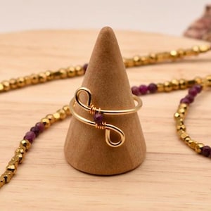 May include: A gold-coloured ring with a wire design and a small purple bead is displayed on a wooden cone. A gold necklace and bracelet with purple beads are also present. The jewellery is set on a wooden surface with a seashell.