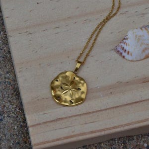 May include: A gold-colored necklace with a sand dollar pendant. The pendant has a textured surface and a star-like design in the center. The necklace is displayed on a wooden surface with seashells.