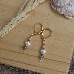 Könnte beinhalten: Goldfarbene Klappcreolen mit Süßwasserperlen, rosa und grünen Perlen. Die Ohrringe werden auf einer Holzoberfläche mit einer Muschel und getrocknetem Gras präsentiert.