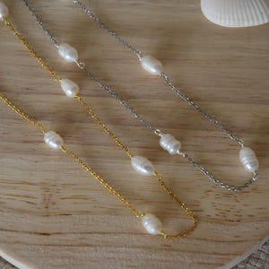 Puede incluir: Tres delicados collares con perlas ovaladas blancas. Uno es dorado, otro plateado y el tercero también plateado. Los collares se exhiben sobre una superficie de madera clara, con una concha marina en el fondo.
