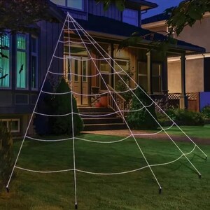 May include: A large white spider web decoration with six support poles, set up in a front garden at night.
