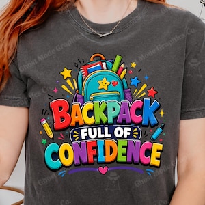 Op de afbeelding: Een antracietgrijs t-shirt met een kleurrijke afbeelding van een blauwgroene rugzak omringd door schoolspullen zoals potloden en sterren, met de tekst "BACKPACK FULL OF CONFIDENCE" in dikke, veelkleurige letters op de borst.