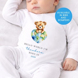 May include: White baby romper featuring a teddy bear on a heart-shaped globe. The text says "Hello world I'm Theodore born in 2026." A blue circle indicates availability in bibs and rompers.