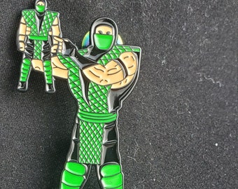 May include: A green and black enamel pin featuring a ninja character holding a smaller version of themself. The ninja is wearing a green mask and a green and black outfit.