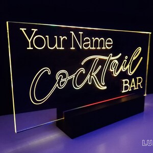 May include: A clear acrylic sign with a black base, illuminated with warm white LED lights. The sign reads "Your Name Cocktail Bar" in a white, cursive font.