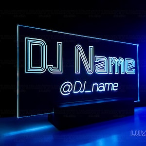 以下が含まれることがあります： 黒い台座に「DJ Name @DJ_name」という文字が書かれた青いネオンサイン。サインは点灯しており、暗い背景に対して文字がはっきり見える。