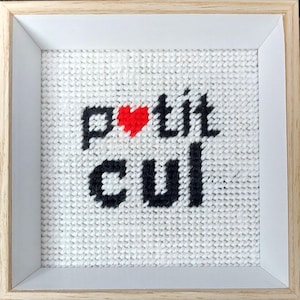 Puede incluir: Obra de arte bordada a punto de cruz enmarcada con las palabras "petit cul" en negro, un corazón rojo reemplaza la "o" en "petit". La obra está sobre un fondo blanco tejido, enmarcada en un marco de madera clara.