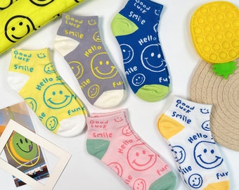 May include: A collection of colorful ankle socks with smiley face designs. The socks are in various colors, including yellow, blue, pink, gray, and white, with the words "Good Luck Smile" and "Hello Fun" printed on them. A yellow smiley face bag and a yellow pop-it toy are also visible.