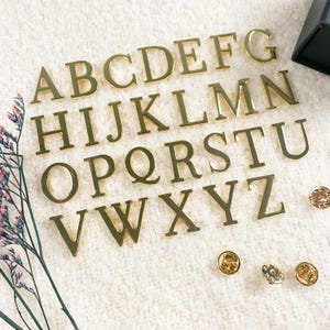 May include: A set of gold-colored metal alphabet letters, A through Z, arranged in alphabetical order. The letters are on a white surface with two gold-colored metal pins.