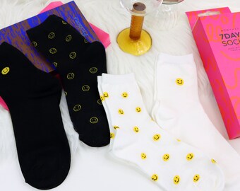 May include: A set of three pairs of socks with a smiley face pattern. One pair is black with yellow smiley faces, one pair is white with yellow smiley faces, and one pair is white with a single yellow smiley face. The socks are packaged in a pink box with the text "7 DAY SOCKS" printed on it.