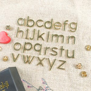 May include: Gold-colored metal alphabet letters, from A to Z, arranged in three rows on a beige fabric surface.