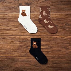 May include: Three pairs of crew socks in white, brown, and black. The white sock features a single brown teddy bear graphic. The brown sock has an all-over pattern of small teddy bears. The black sock displays a single brown teddy bear graphic.