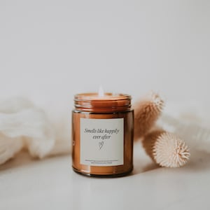 May include: A brown glass candle jar with a white label that reads "Smells like happily ever after" with a small heart symbol. The candle is lit and the flame is visible.