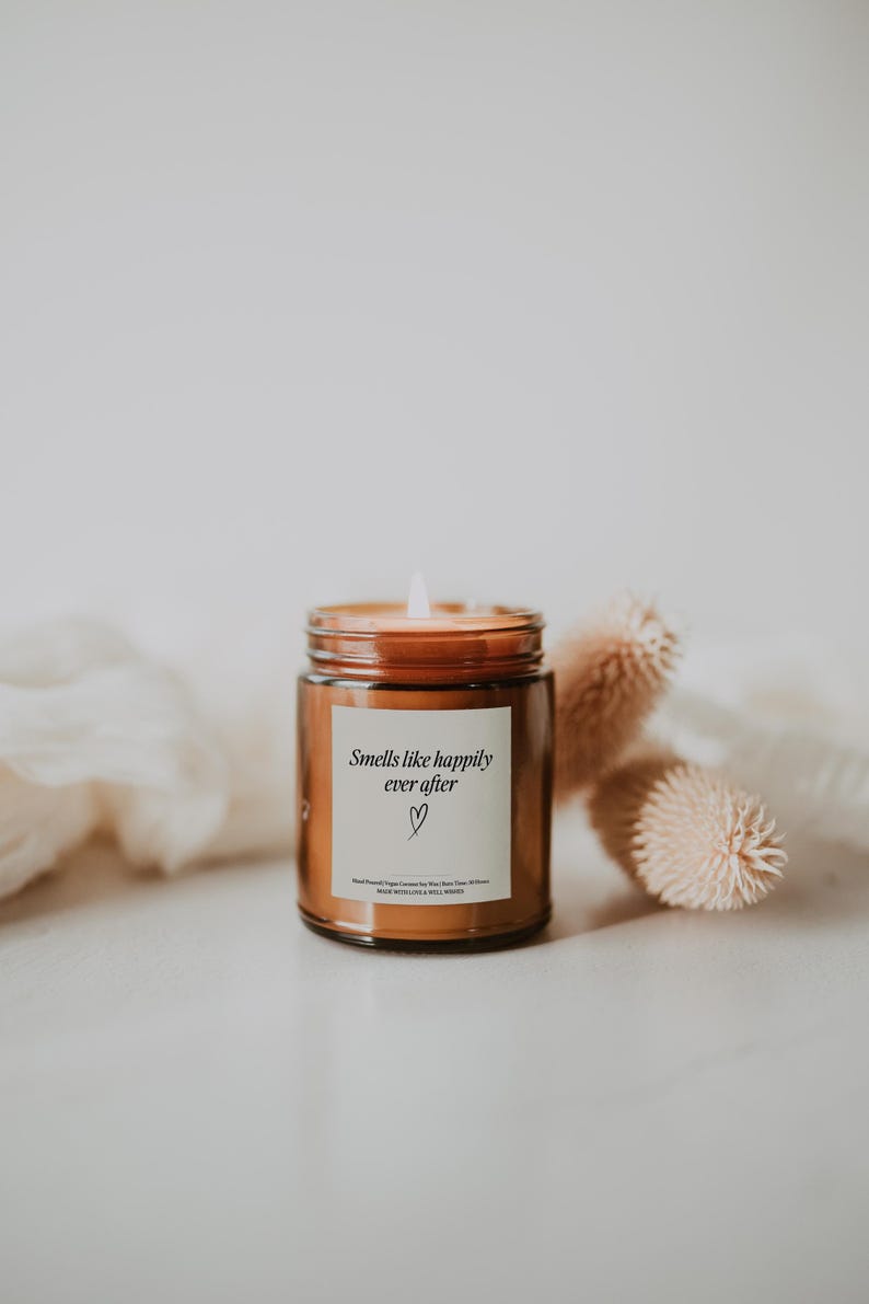 May include: A brown glass candle jar with a white label that reads "Smells like happily ever after" with a small heart symbol. The candle is lit and the flame is visible.