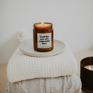 May include: A brown glass candle with a white label that reads "Smells like your cat's judgmental stare." The candle is sitting on a white plate on top of a stack of white knitted blankets.