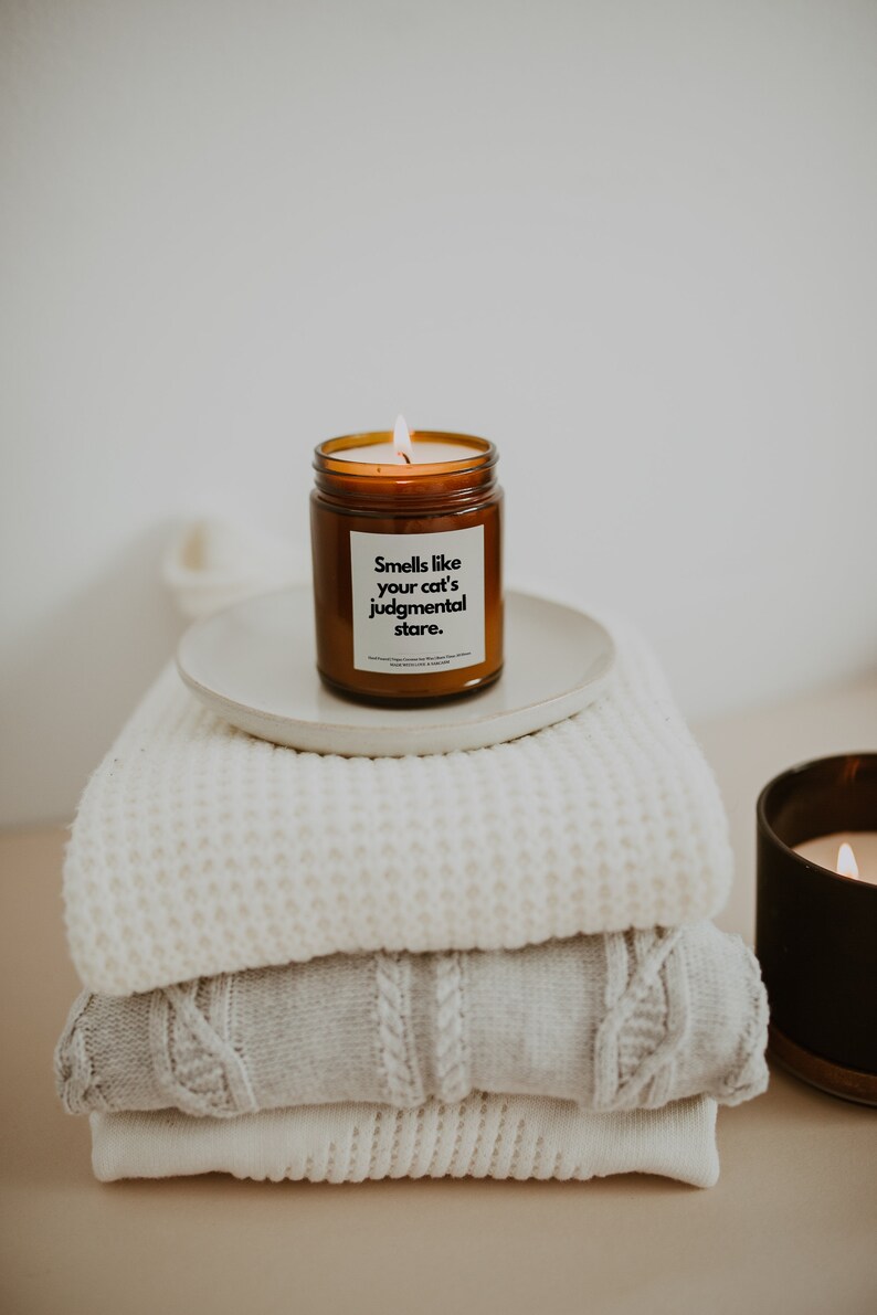 May include: A brown glass candle with a white label that reads "Smells like your cat's judgmental stare." The candle is sitting on a white plate on top of a stack of white knitted blankets.