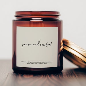 May include: A brown glass candle jar with a gold lid. The label reads "peace and comfort" and includes the text "Hand Poured | Vegan Coconut Soy Wax | Burn Time: 50 Hours". The label also says "MADE WITH LOVE & CONDOLENCES".