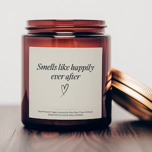 May include: A brown glass candle jar with a gold lid. The label reads "Smells like happily ever after" with a heart symbol. The candle is hand-poured with vegan coconut soy wax and has a burn time of 50 hours.