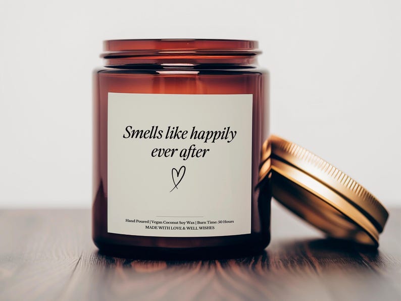 May include: A brown glass candle jar with a gold lid. The label reads "Smells like happily ever after" with a heart symbol. The candle is hand-poured with vegan coconut soy wax and has a burn time of 50 hours.